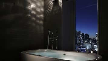 A modern bathroom with a bathtub, a city view through a large window, and a wall-mounted light fixture.
