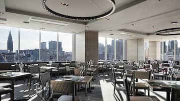 A modern restaurant with a city skyline view, featuring round tables, comfortable seating, and a circular ceiling light fixture.
