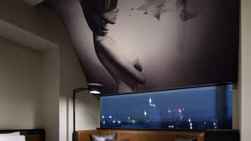 A hotel room with a large window, a bed, a sofa, a table, and a wall-mounted TV.