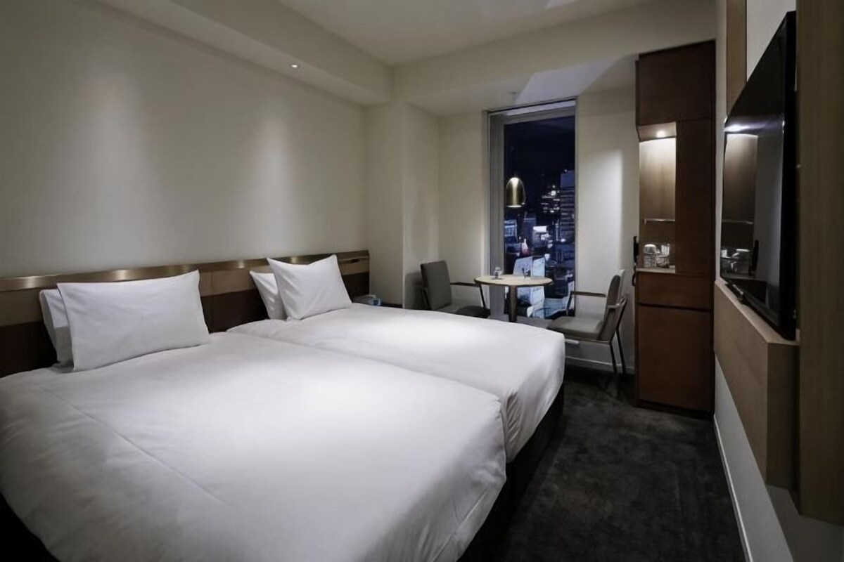 Quarto de hotel com duas camas, uma escrivaninha, uma cadeira, uma TV e vista para a cidade à noite.