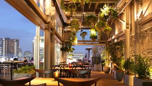 An indoor dining area with tables and chairs, hanging plants, and a city view through large windows. - Shinjuku Granbell Hotel (Tokyo)