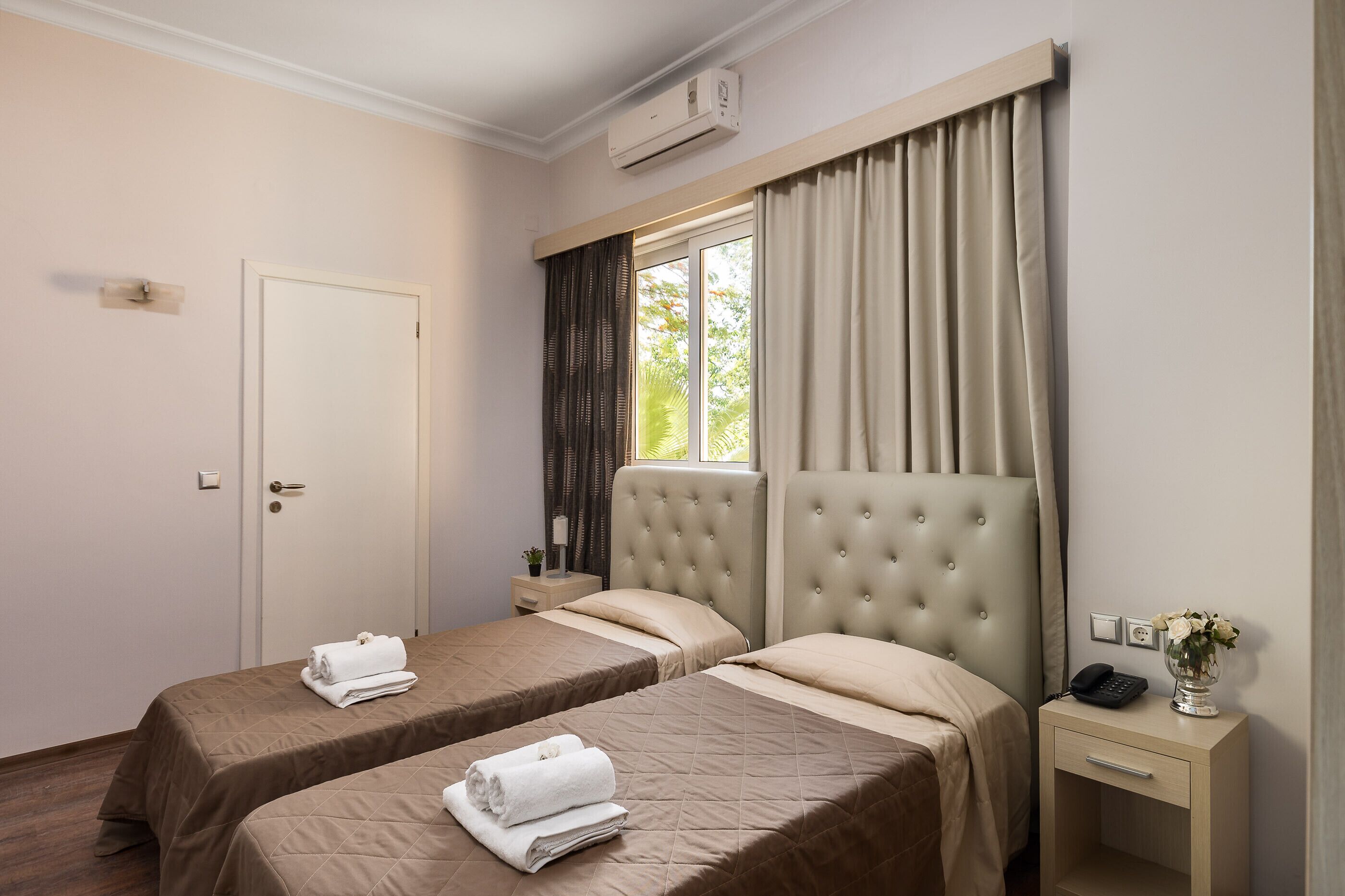Hotellihuone, jossa on kaksi sänkyä, yöpöytä lampun kanssa, ikkuna verhoilla ja ilmastointilaite.
