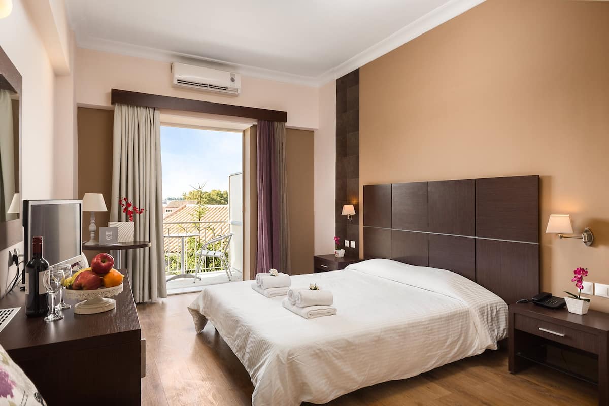 Une chambre d’hôtel avec un grand lit, un bureau avec un ordinateur et un panier de fruits, une table de chevet avec une lampe, et une vue sur l’extérieur.