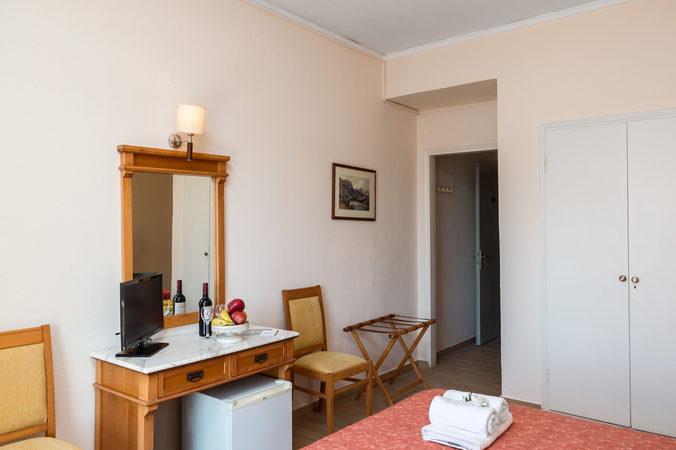 A hotel room with a wooden desk, a mirror, a television, a small refrigerator, a chair, a bed with a red bedspread, a drying rack, and a wardrobe.