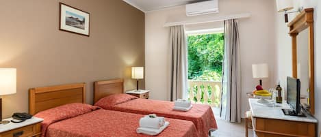 A hotel room with two beds, a desk with a computer, a TV, and a balcony with a view of greenery.