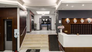 Zona de recepción de un hotel con una escalera que sube al piso superior, un mostrador de recepción y una pared con varios relojes.
