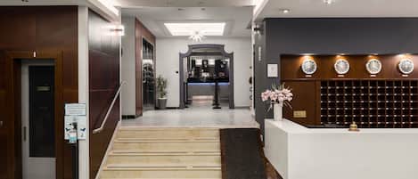 A hotel reception area with a staircase leading to the upper floor, a reception desk, and a wall with multiple clocks.