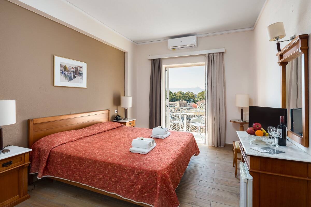 Une chambre d’hôtel avec un grand lit, un bureau avec un miroir, une télévision et un balcon offrant une vue sur les bâtiments et les arbres.