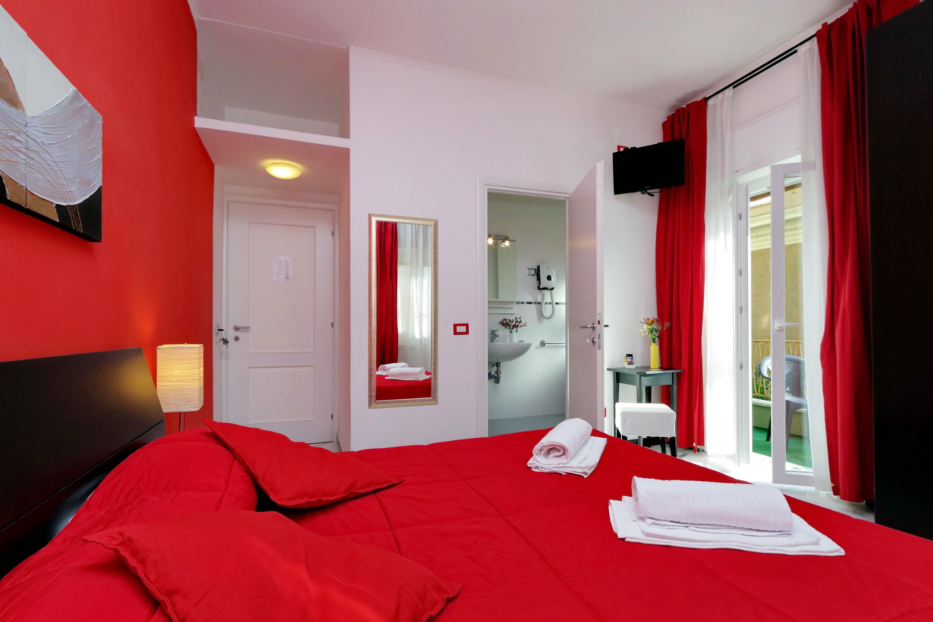 A hotel room with a red bedspread, a white door, a bathroom visible through an open door, and a balcony with red curtains.