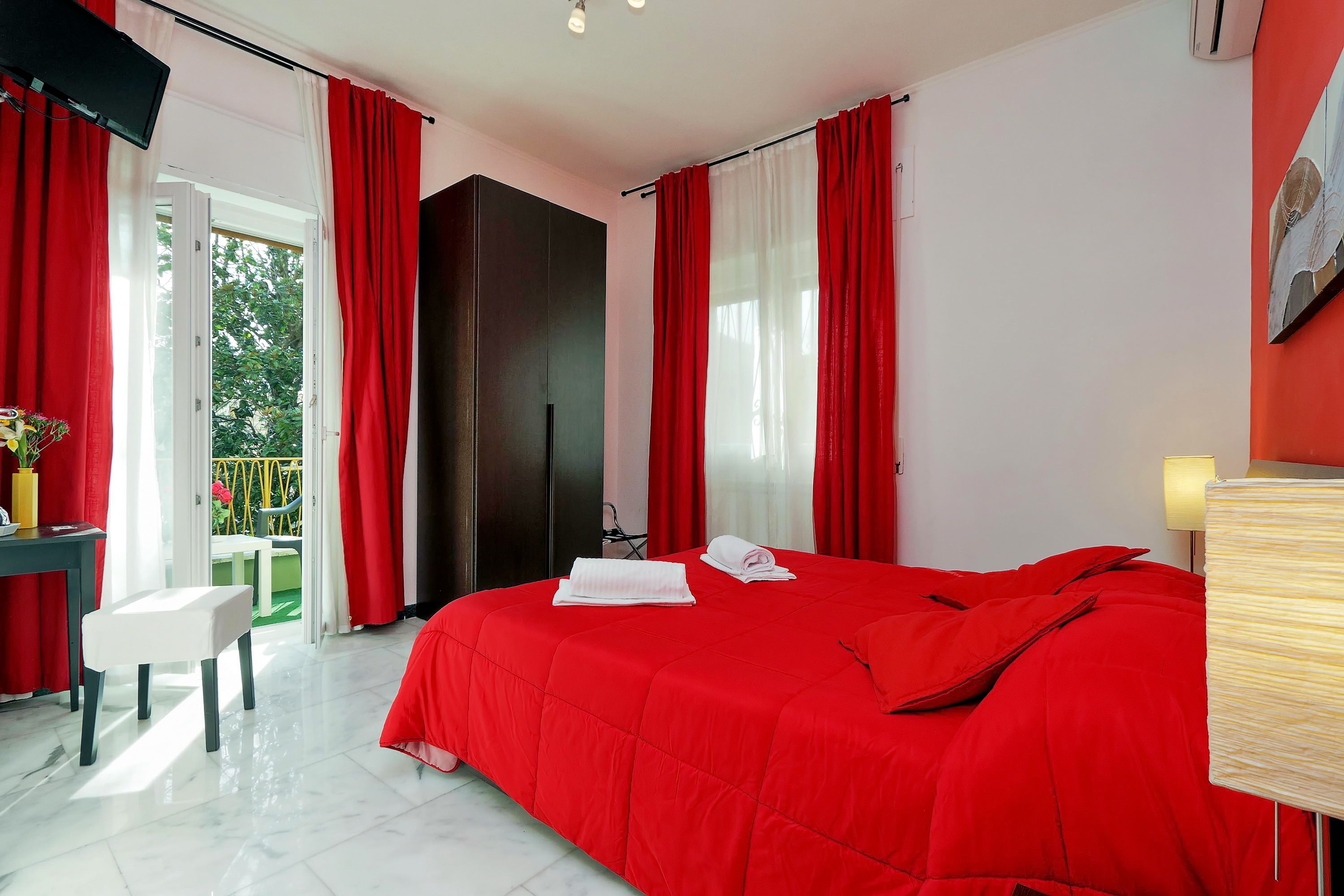 A hotel room with a large bed, red curtains, a TV, and a balcony with a view of trees.