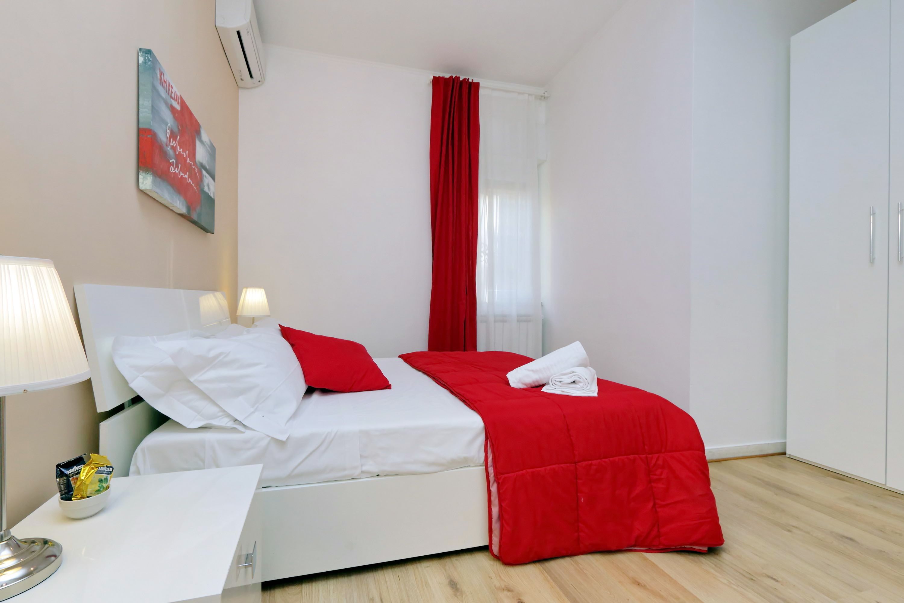 A hotel room with a bed, a bedside table with a lamp, a red curtain, and a white wardrobe.
