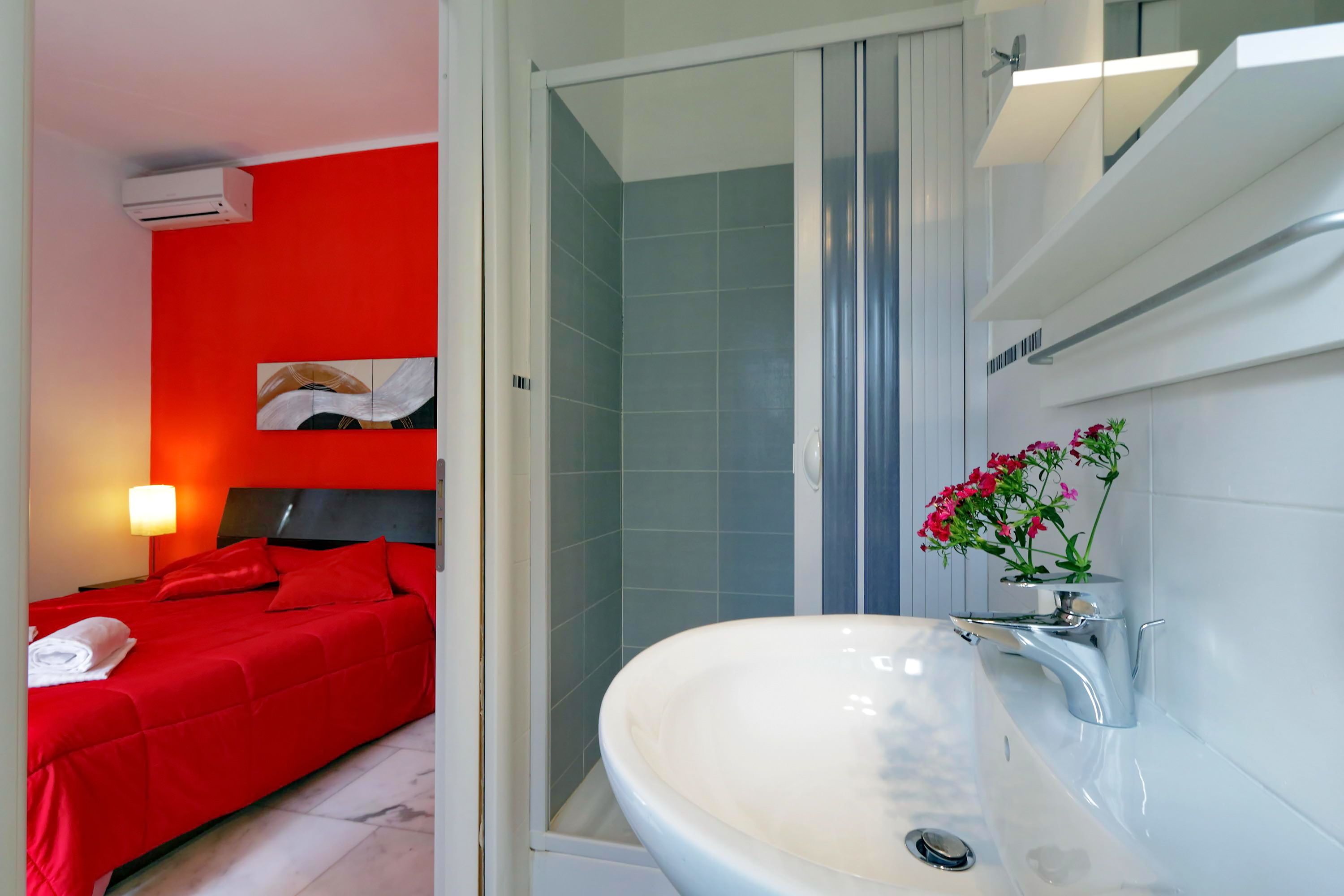 A hotel room with a red bed, a white sink with a mirror, and a shower area.