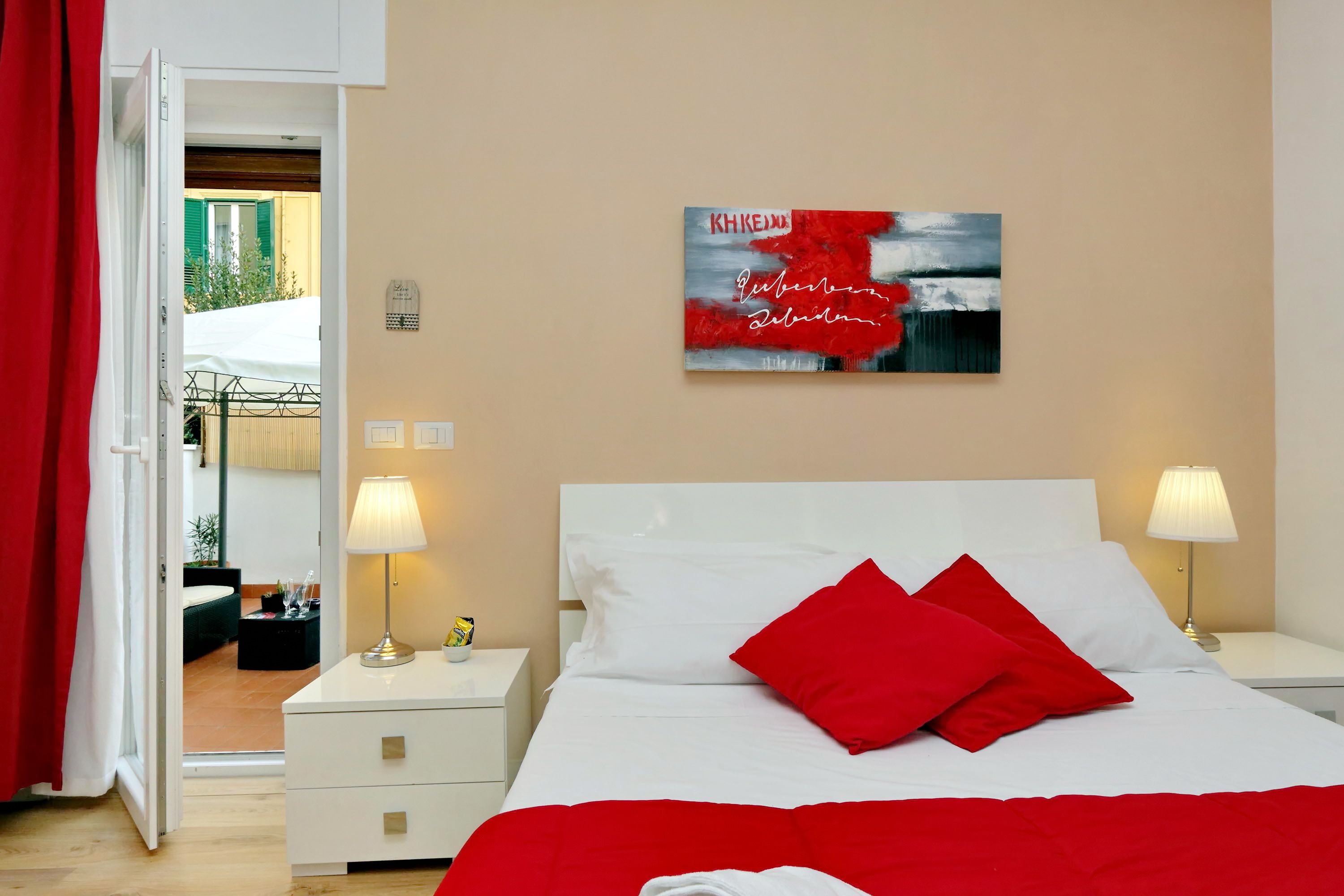 A hotel room with a bed, two bedside lamps, a red throw blanket, and a painting on the wall.