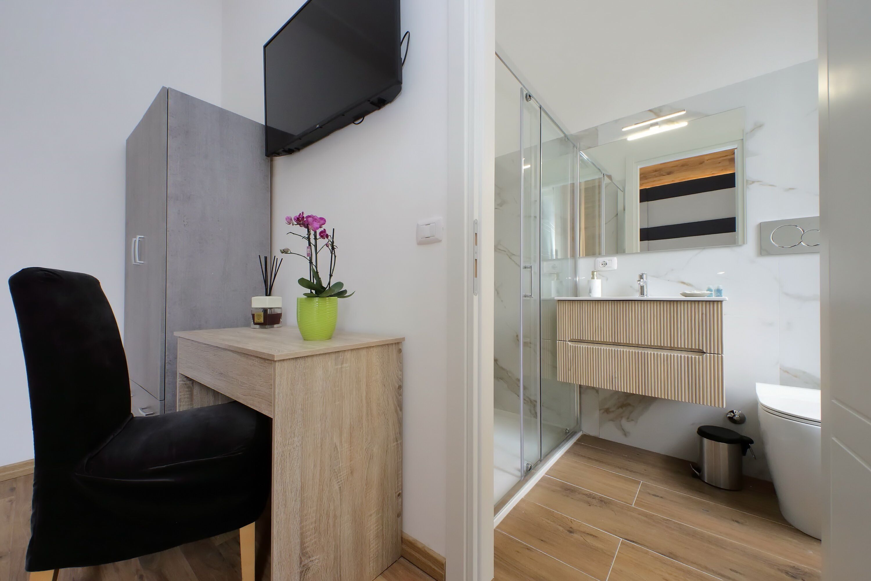 A modern bathroom with a glass shower enclosure, a wooden vanity, a black chair, and a mounted television.