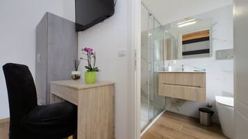 A modern bathroom with a glass shower enclosure, a wooden vanity, a black chair, and a mounted television.