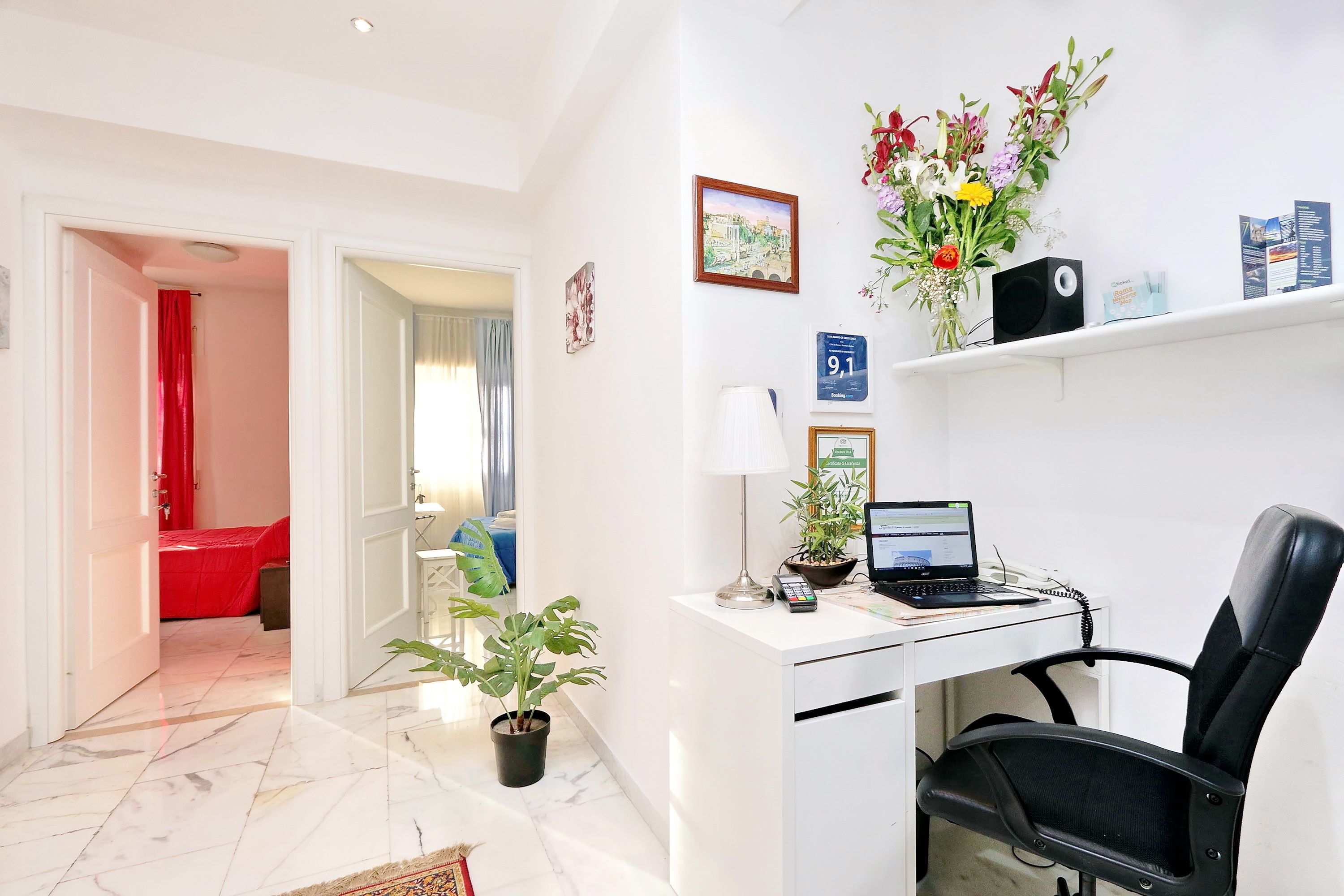 A hotel reception area with a desk, computer, chair, potted plants, and a red bed in the background.