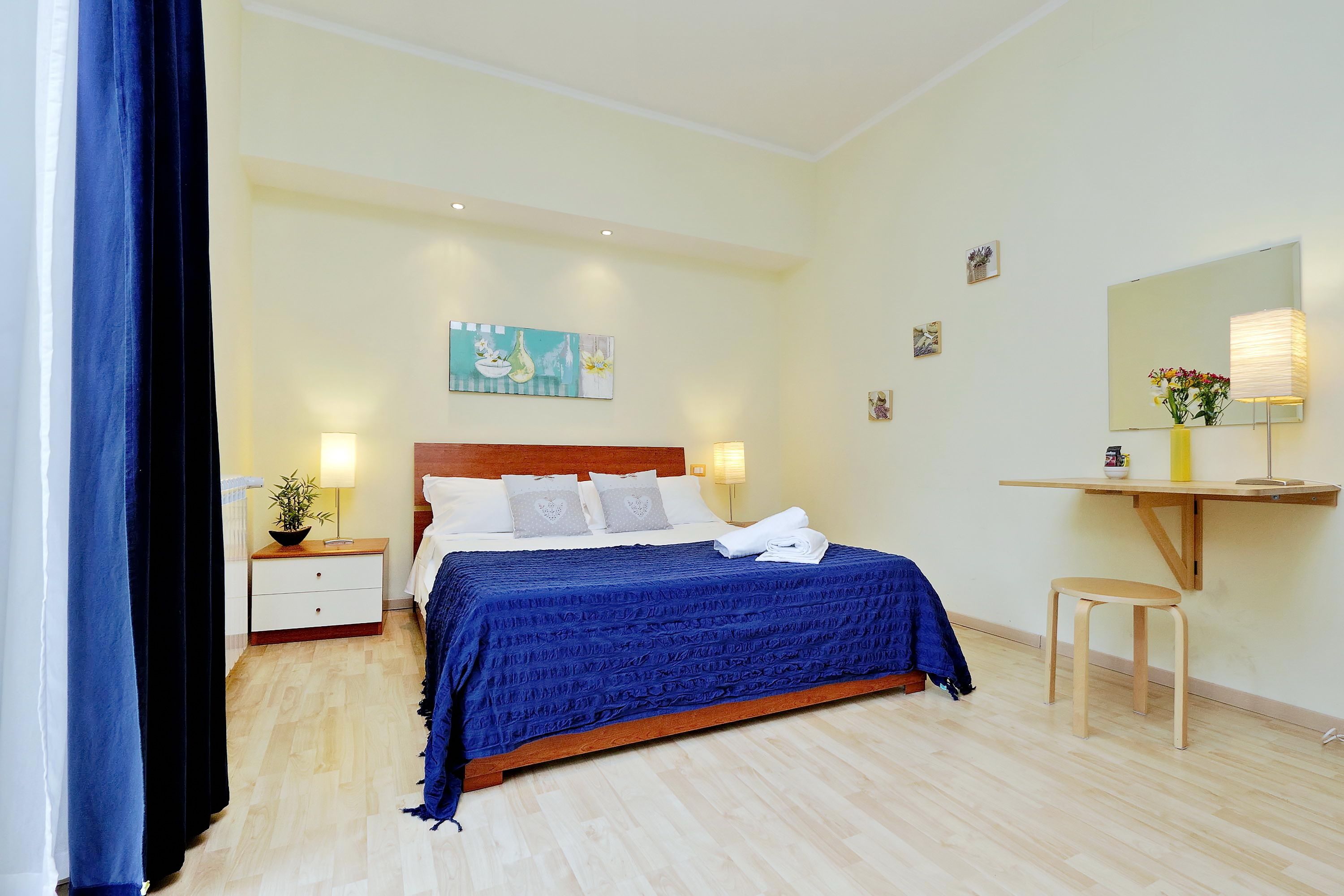 A bedroom with a bed, bedside tables, a nightstand, a desk, a chair, a mirror, and artwork on the walls.