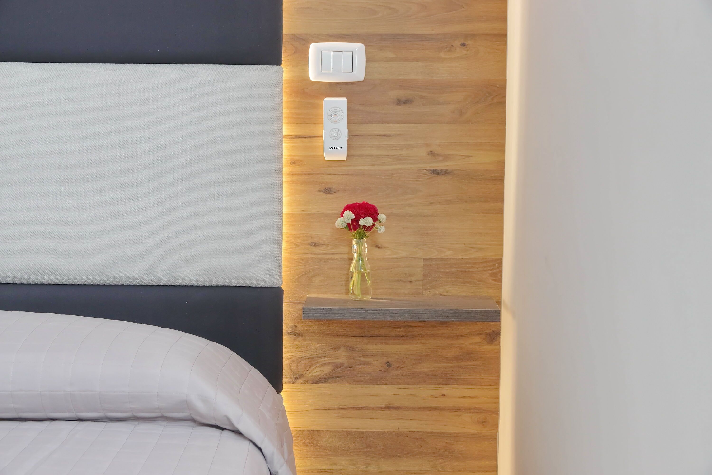 A bedroom with a bed, a wooden nightstand, a wall-mounted light switch, and a small vase with flowers.