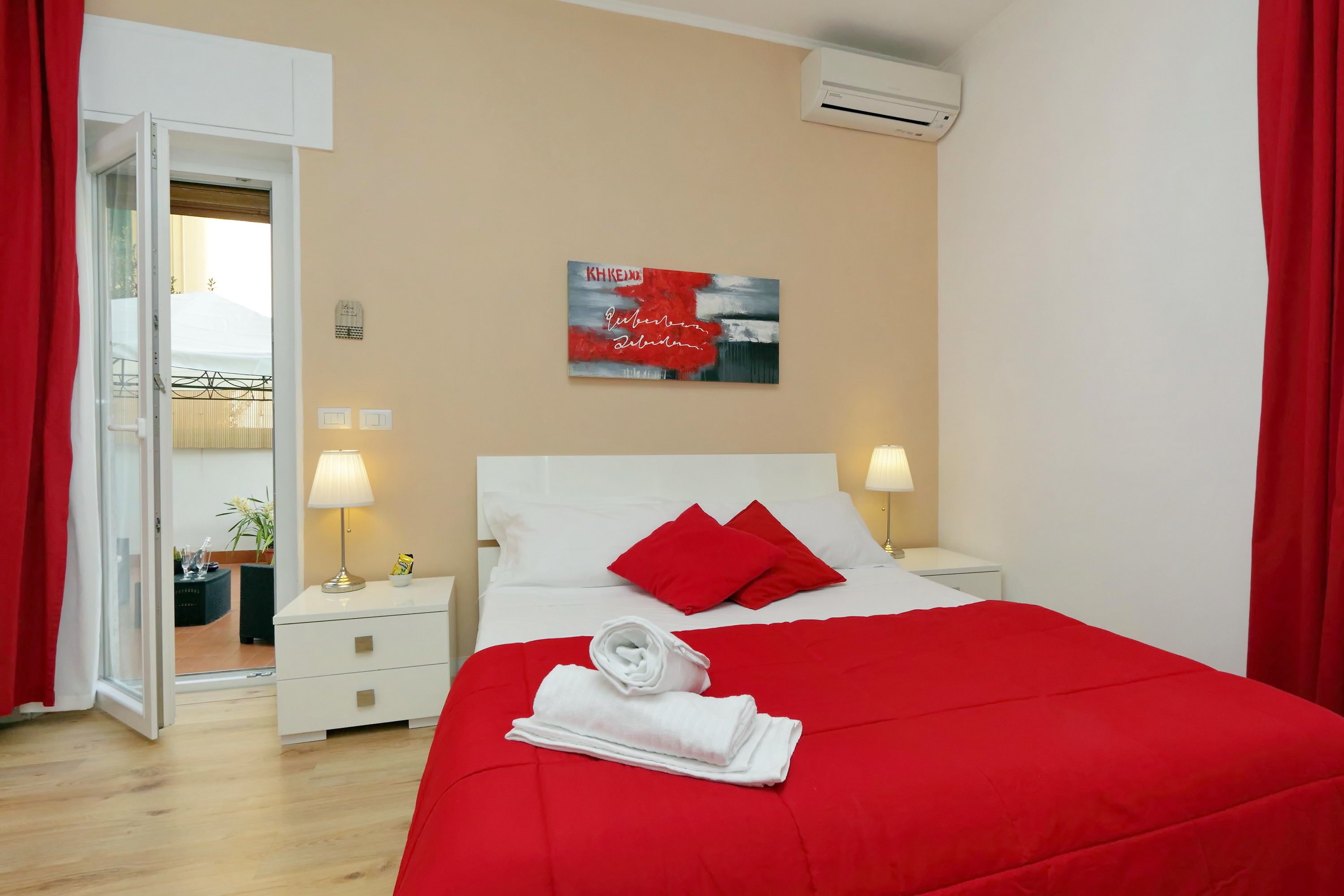 A hotel room with a bed, red curtains, a wall-mounted air conditioner, a bedside lamp, and a painting.