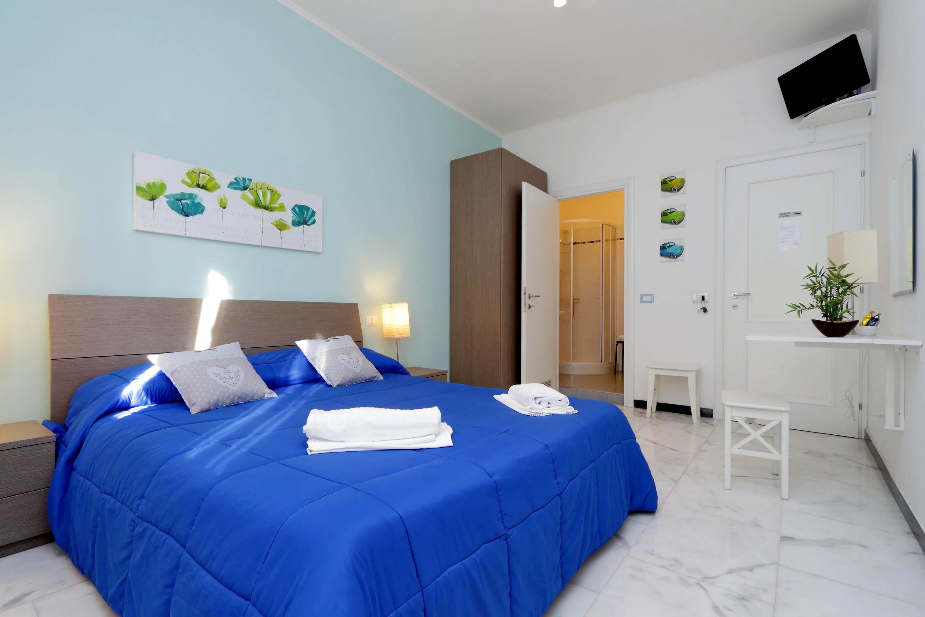 Camera d'albergo con un letto grande, un comodino, una TV a parete, un tavolino con una pianta e una porta che dà sul bagno.