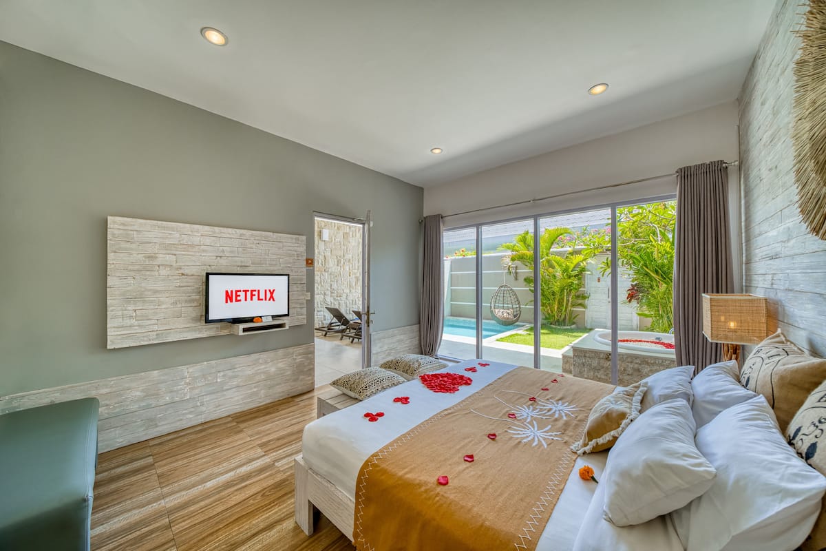 A modern bedroom with a large bed, a TV mounted on the wall, and a view of a swimming pool and greenery outside.