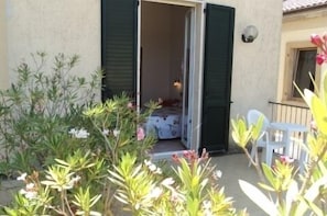 Une pièce avec un lit, une fenêtre et une porte. Il y a une chaise et une table à l’extérieur de la pièce.