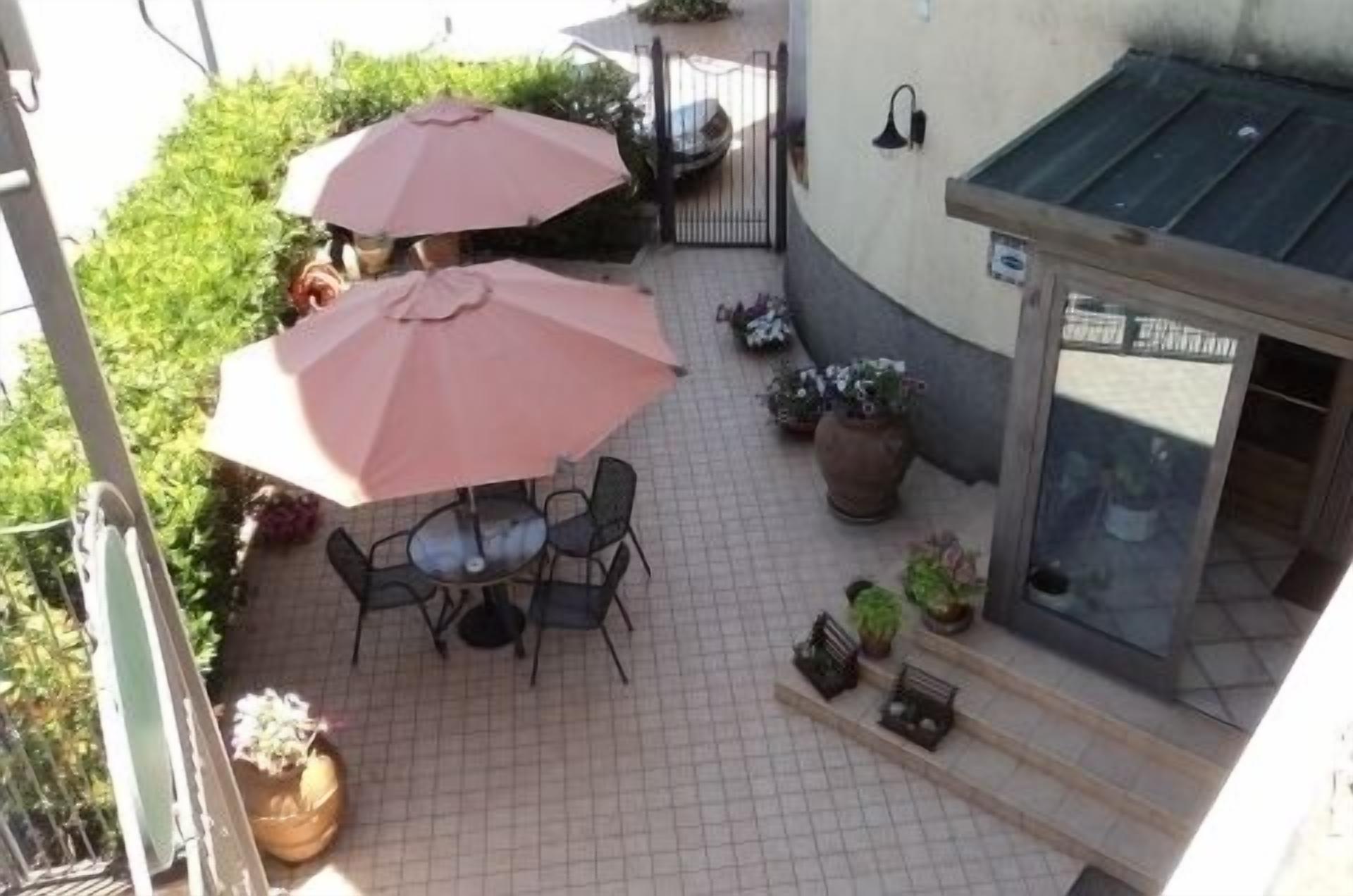 An outdoor patio with two umbrellas, a table, and chairs. There are potted plants and a small garden area.