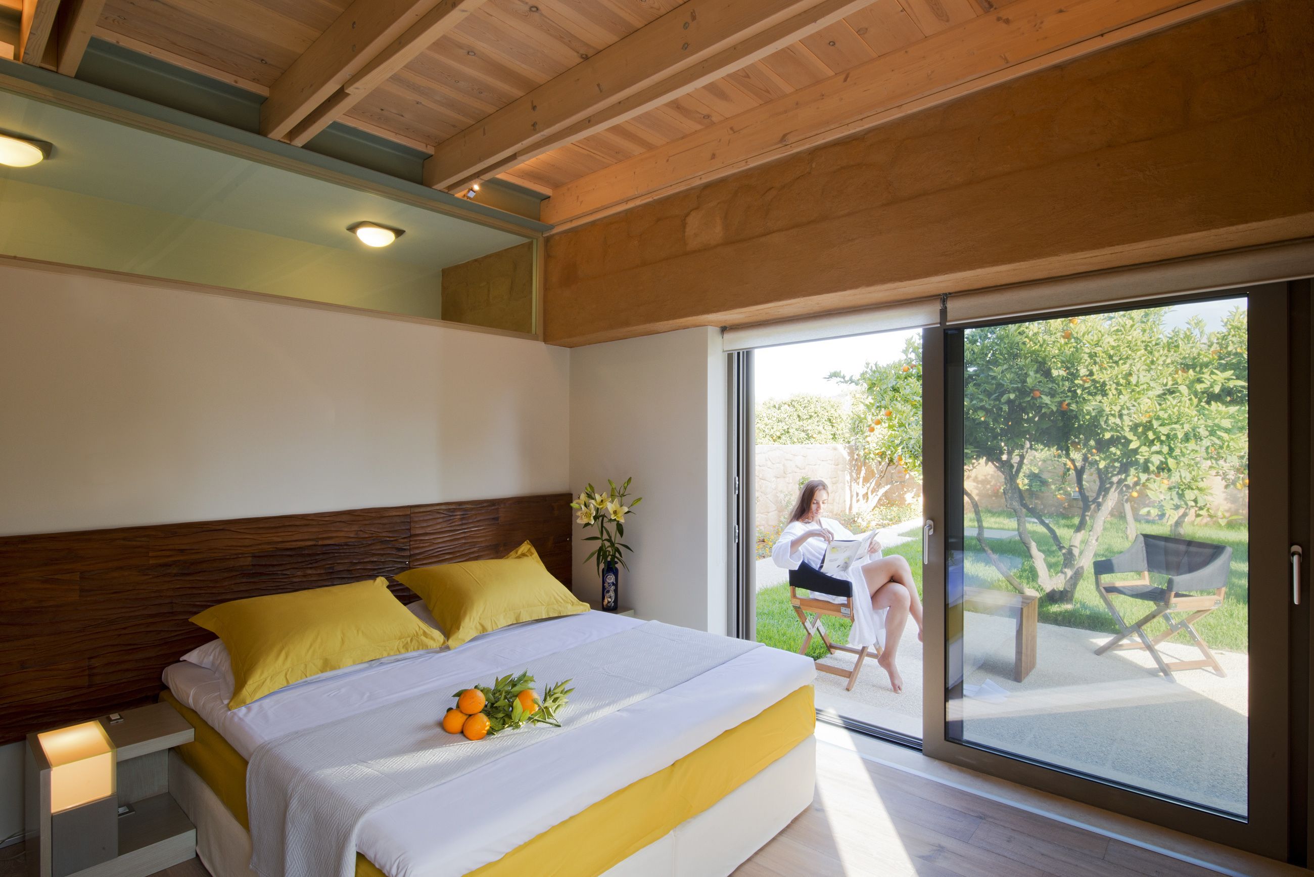 A modern bedroom with a large bed, yellow pillows, and a view of a garden through a sliding glass door.