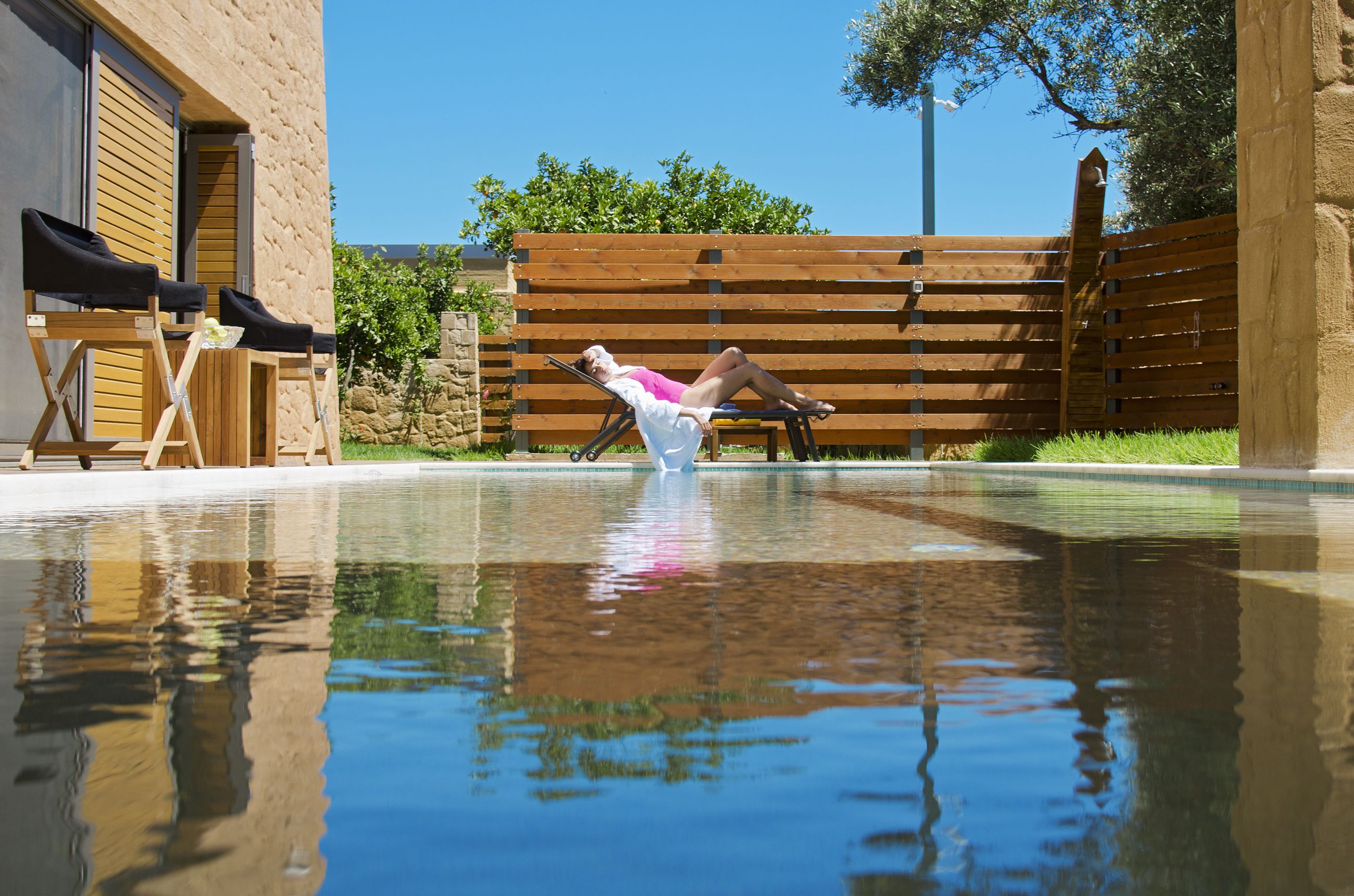 Une zone autour d’une piscine, avec une personne allongée sur une chaise longue, une clôture en bois et un bâtiment doté d’une façade en pierre.