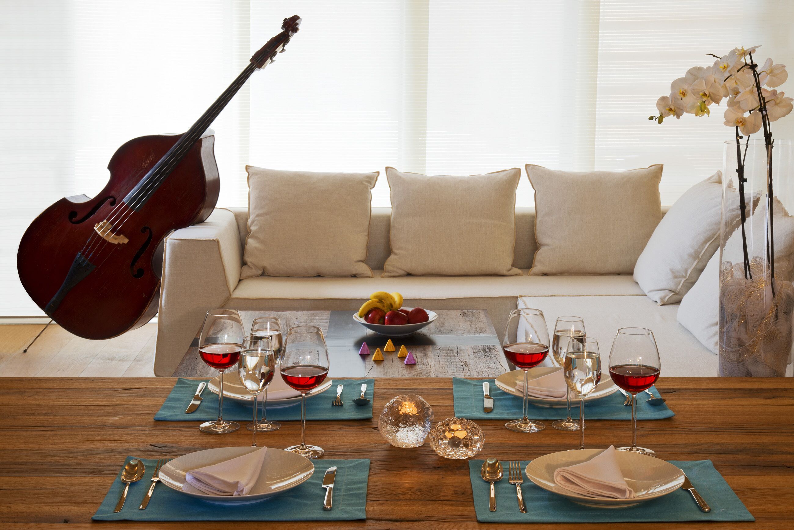 Un comedor con una mesa de madera dispuesta para dos personas, una guitarra apoyada en un sofá y un jarrón con orquídeas.