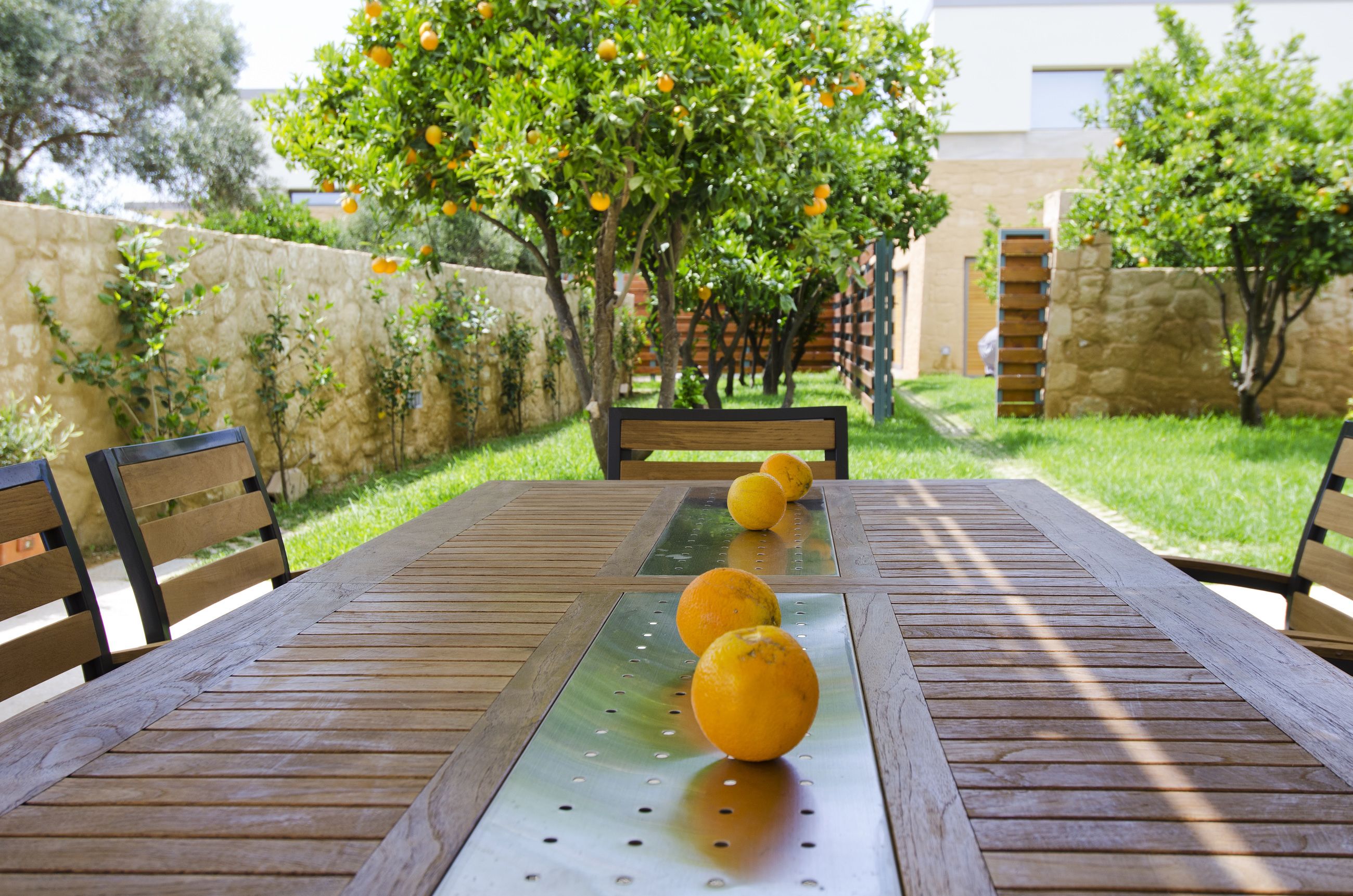 Ein Holztisch mit einem Metalltablett und drei Orangen darauf, draußen mit einer Steinmauer und Bäumen im Hintergrund.