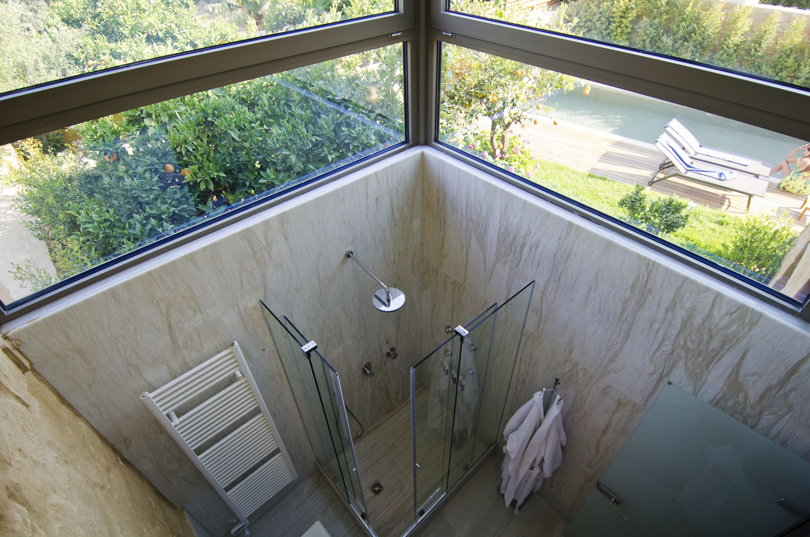A bathroom with a glass-enclosed shower, a towel rack, and a view of a garden through large windows.