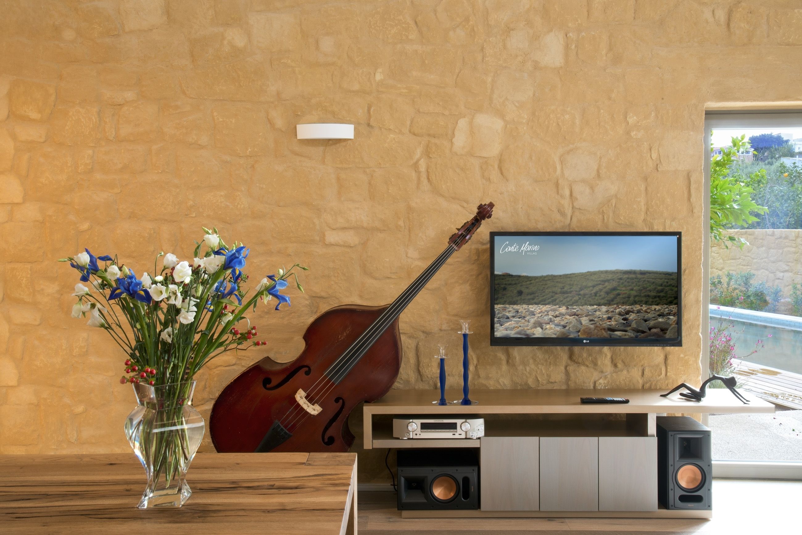 A room with a stone wall, a large TV, a violin, a vase with flowers, and a wooden table.