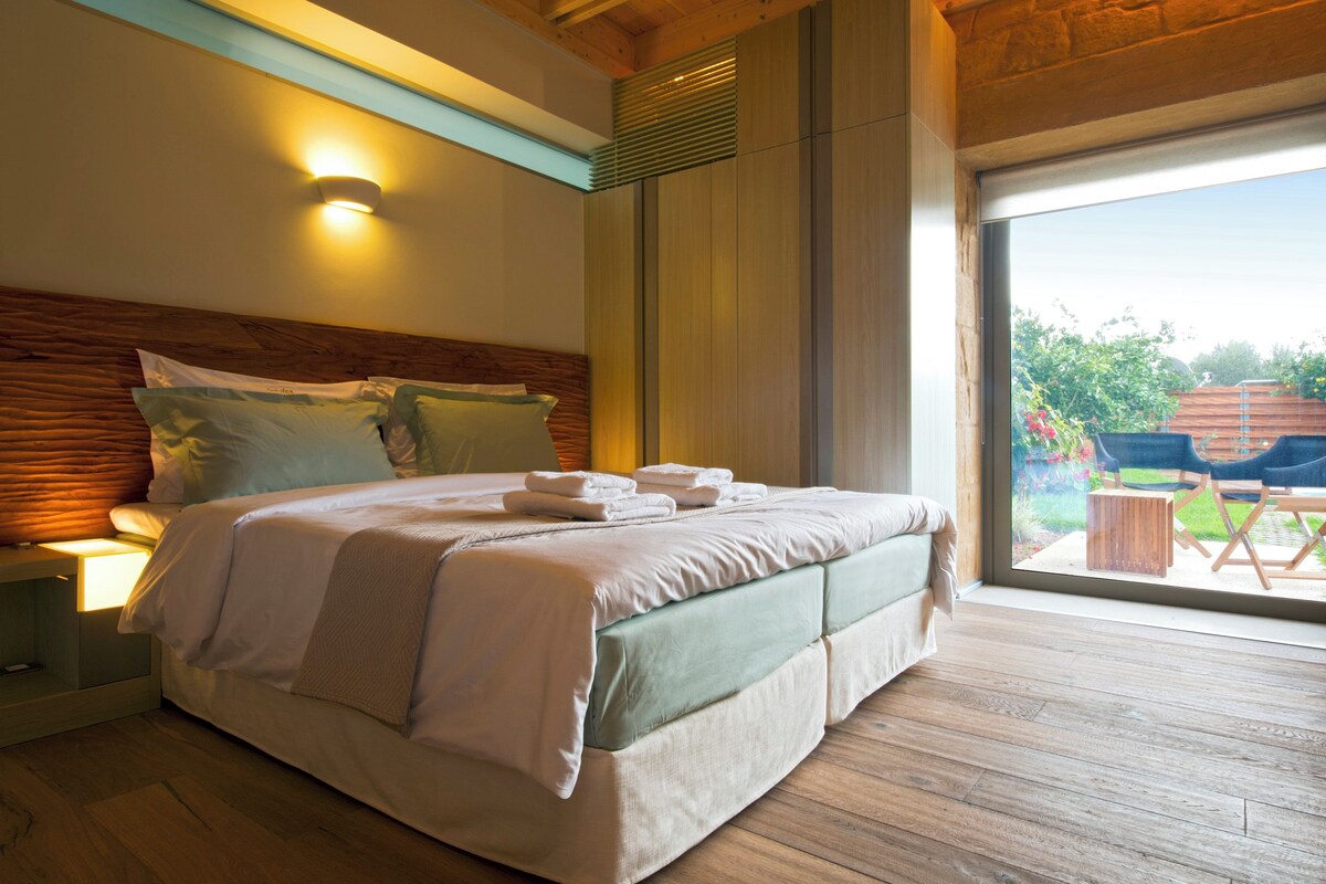 A modern bedroom with a wooden headboard, a large bed with neatly folded towels, and a view of the outdoors through a sliding glass door.