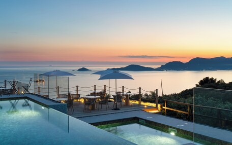 An infinity pool with a view of the sea and mountains at sunset.