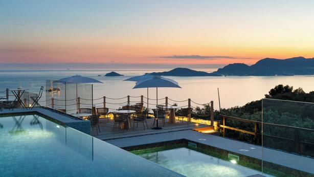 Piscina infinita com vista para o mar e montanhas ao pôr do sol.