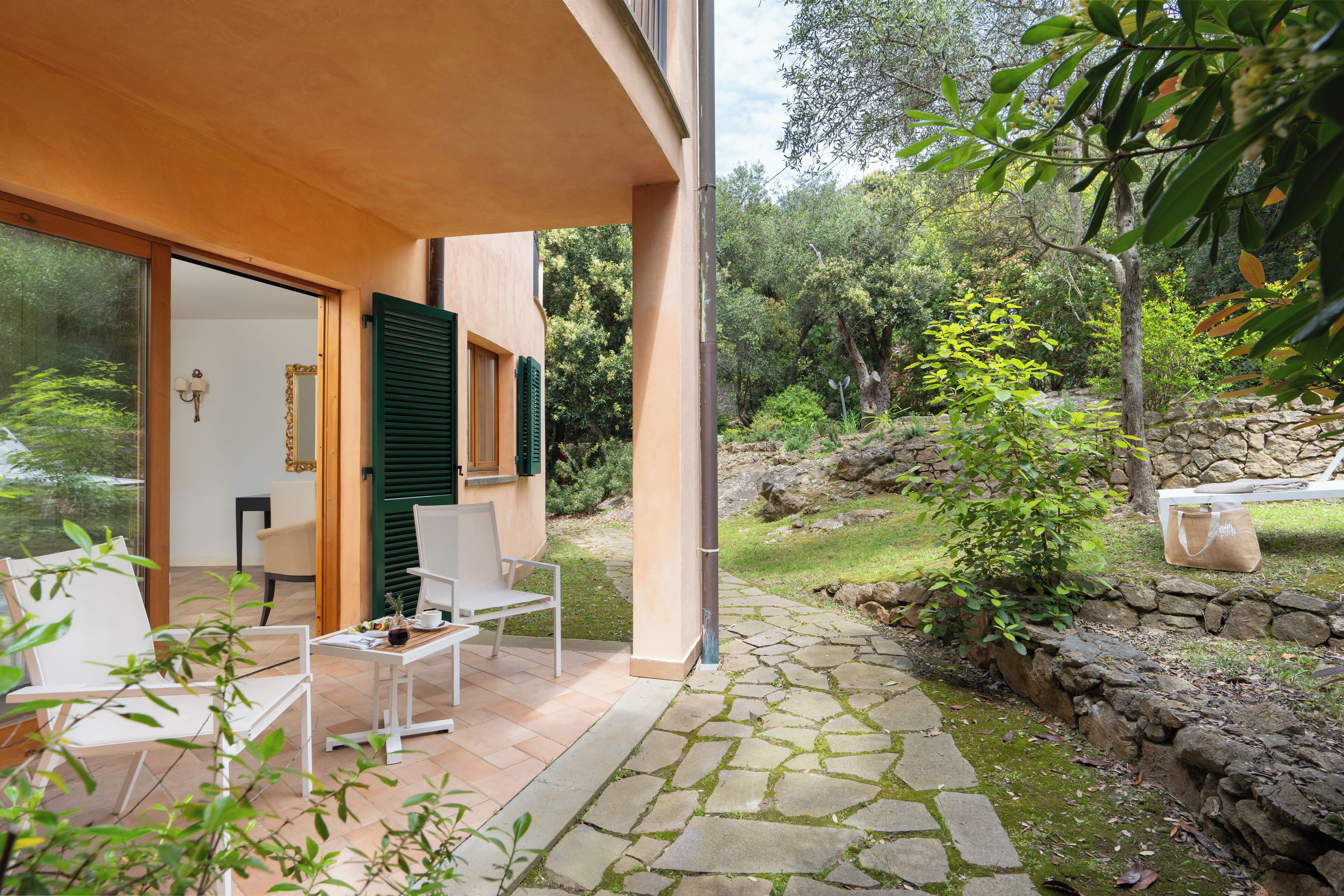 A modern house with a patio area featuring white chairs and a table, surrounded by a stone pathway and greenery.