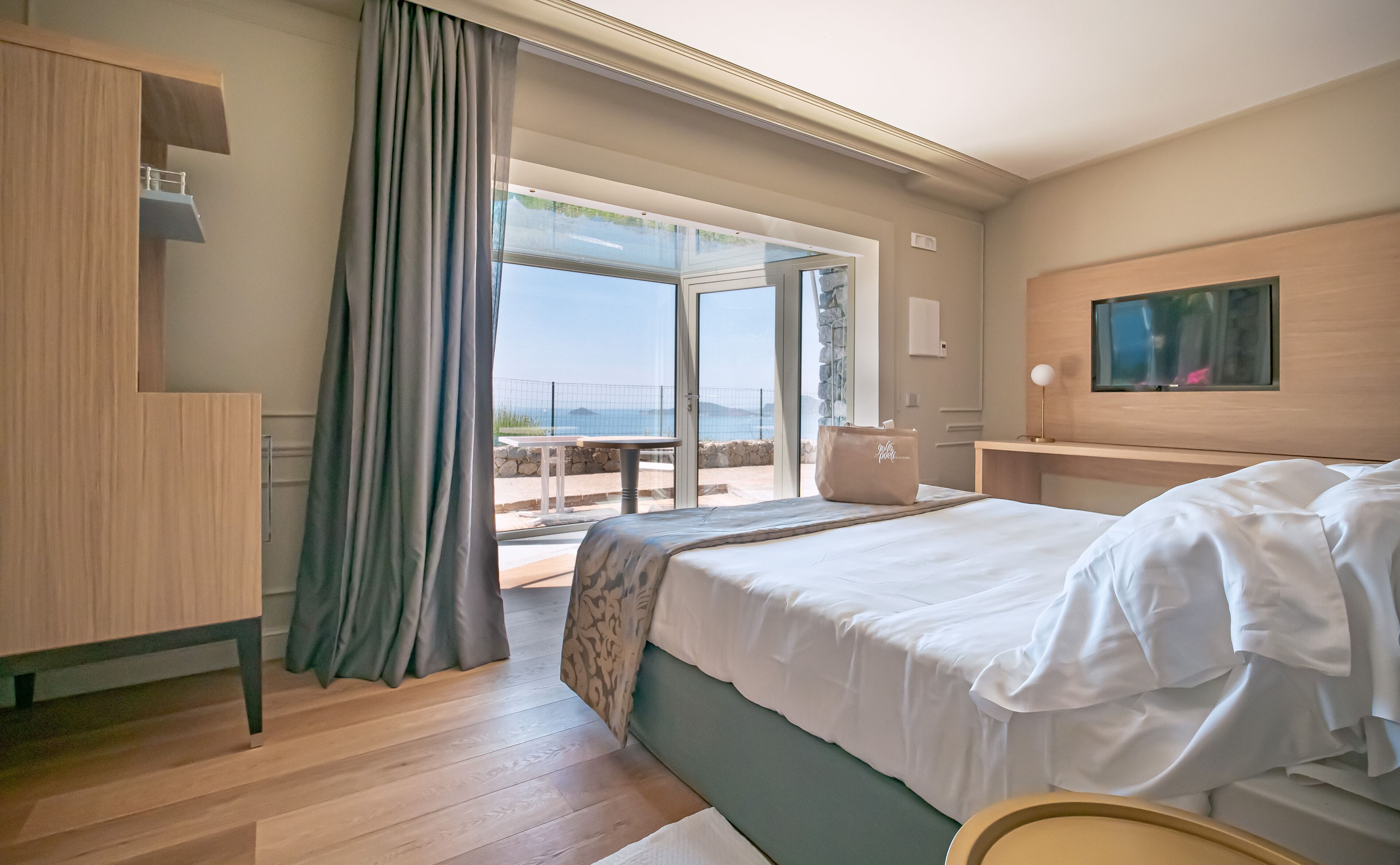A hotel room with a bed, a desk, a chair, a TV, and a balcony with a view of the sea.