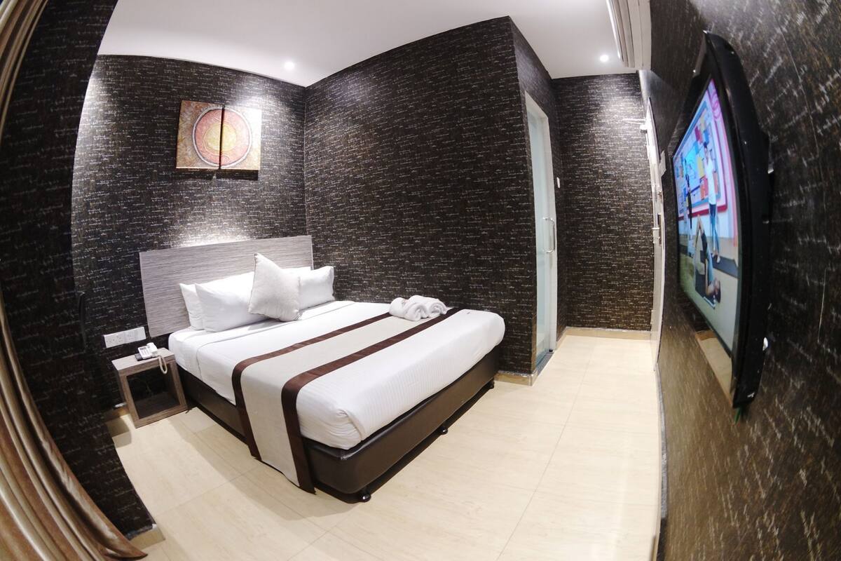 A modern hotel room with a large bed, a nightstand, a TV, and dark textured walls.