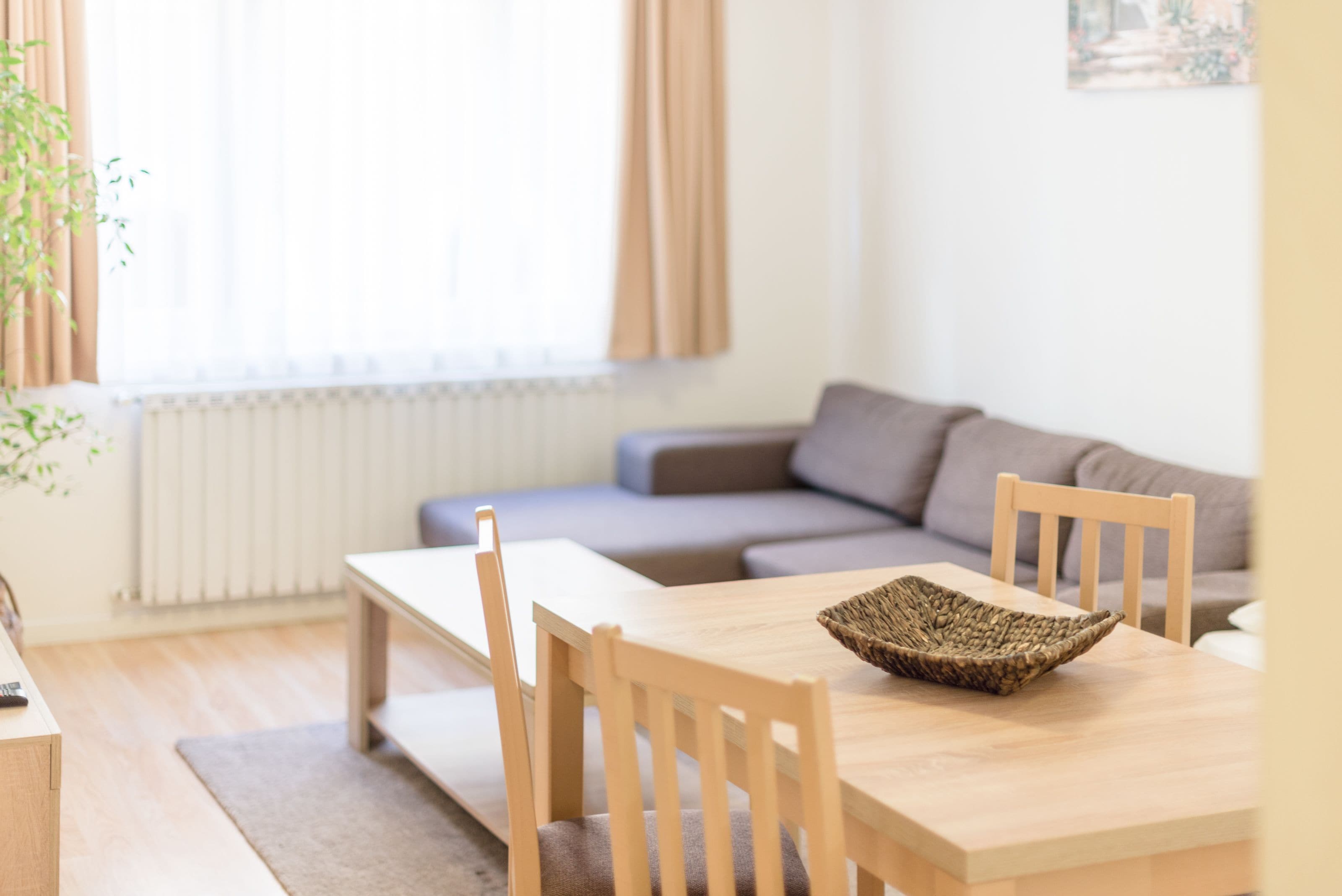 A modern living room with a wooden dining table, two chairs, a grey sofa, and a window with curtains.
