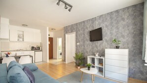 A modern living room with a sofa, a small table, and a TV mounted on the wall. There is a kitchen area in the background with white cabinets and a dining table. - Prince Apartments (Budapest)
