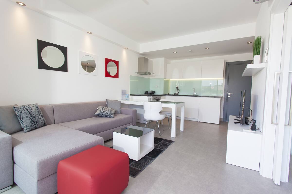 Uma sala de estar moderna com um sofá cinza, um banquinho vermelho e uma mesa de centro de vidro. Há uma área de cozinha com armários brancos e uma mesa de jantar.