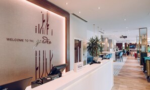 A hotel reception desk with a 'Welcome to the Garden' sign, a digital screen, and a seating area with a bar in the background.