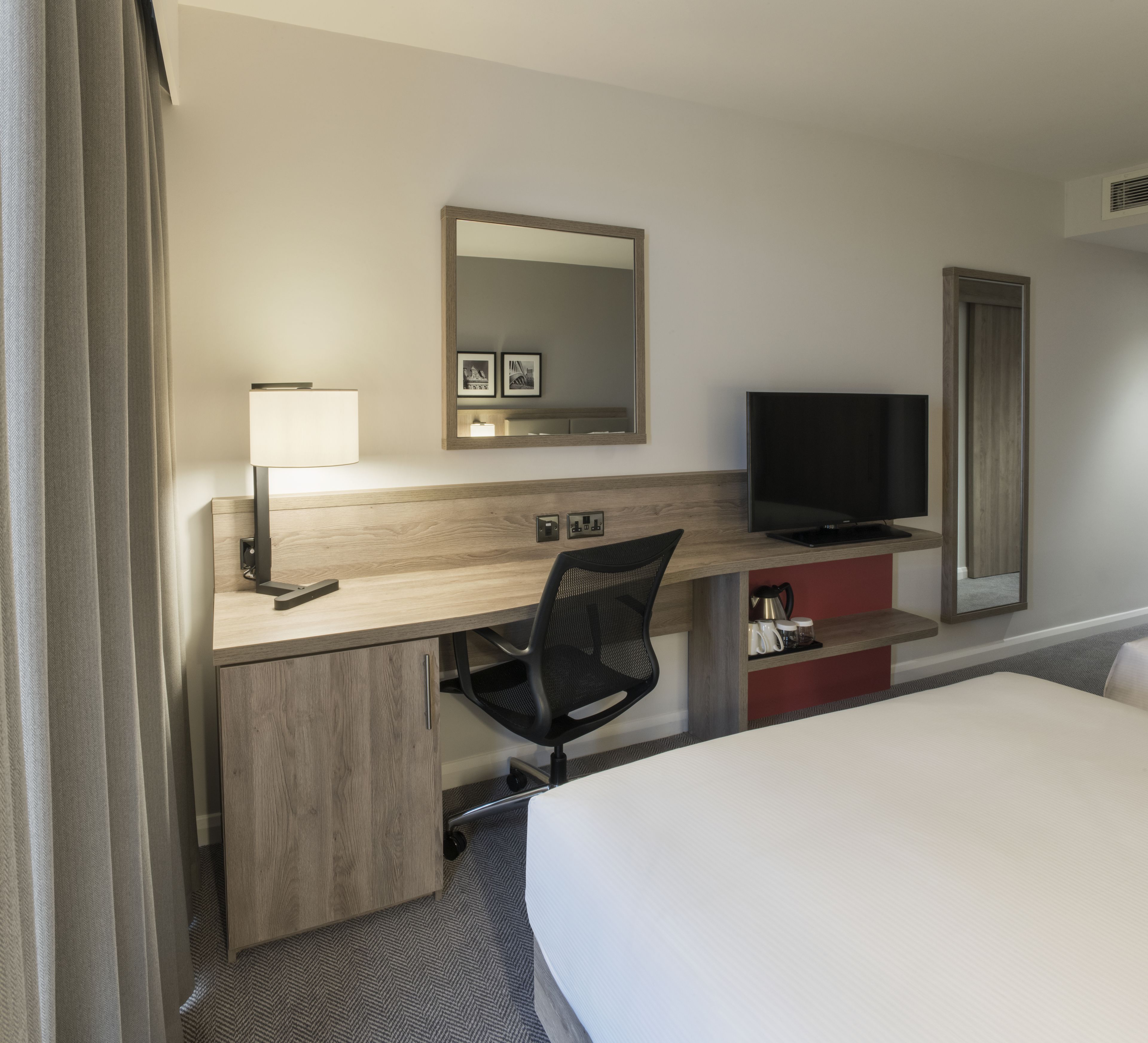 A hotel room with a bed, a desk with a chair, a television, a mirror, and a small shelf with mugs.