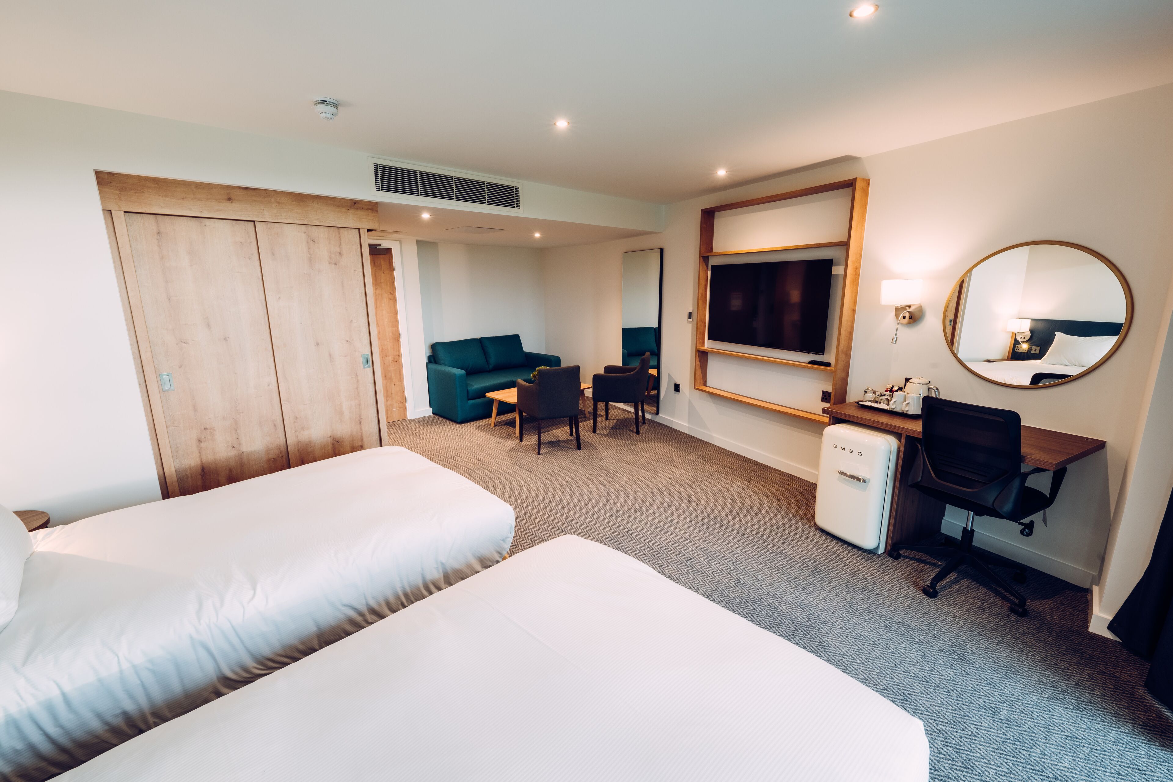 Ein Hotelzimmer mit zwei Betten, einem Schreibtisch, einem Sessel, einer Couch, einem Spiegel und einem Kleiderschrank.
