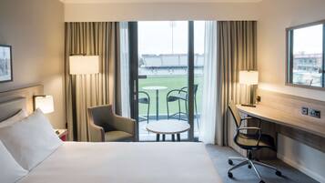 Habitación de hotel con cama, dos sillas, un escritorio y vistas a un campo deportivo a través de amplios ventanales.