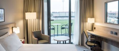 A hotel room with a bed, two chairs, a desk, and a view of a sports field through large windows.