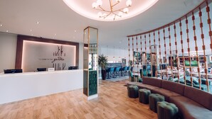 A modern hotel reception area with a wooden reception desk, a seating area with a sofa and ottomans, and a large mirror. - Hilton Garden Inn Manchester Emirates Old Trafford (Manchester)