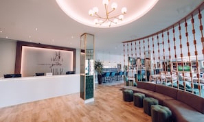 A modern hotel reception area with a wooden reception desk, a seating area with a sofa and ottomans, and a large mirror. - Hilton Garden Inn Manchester Emirates Old Trafford (Manchester)