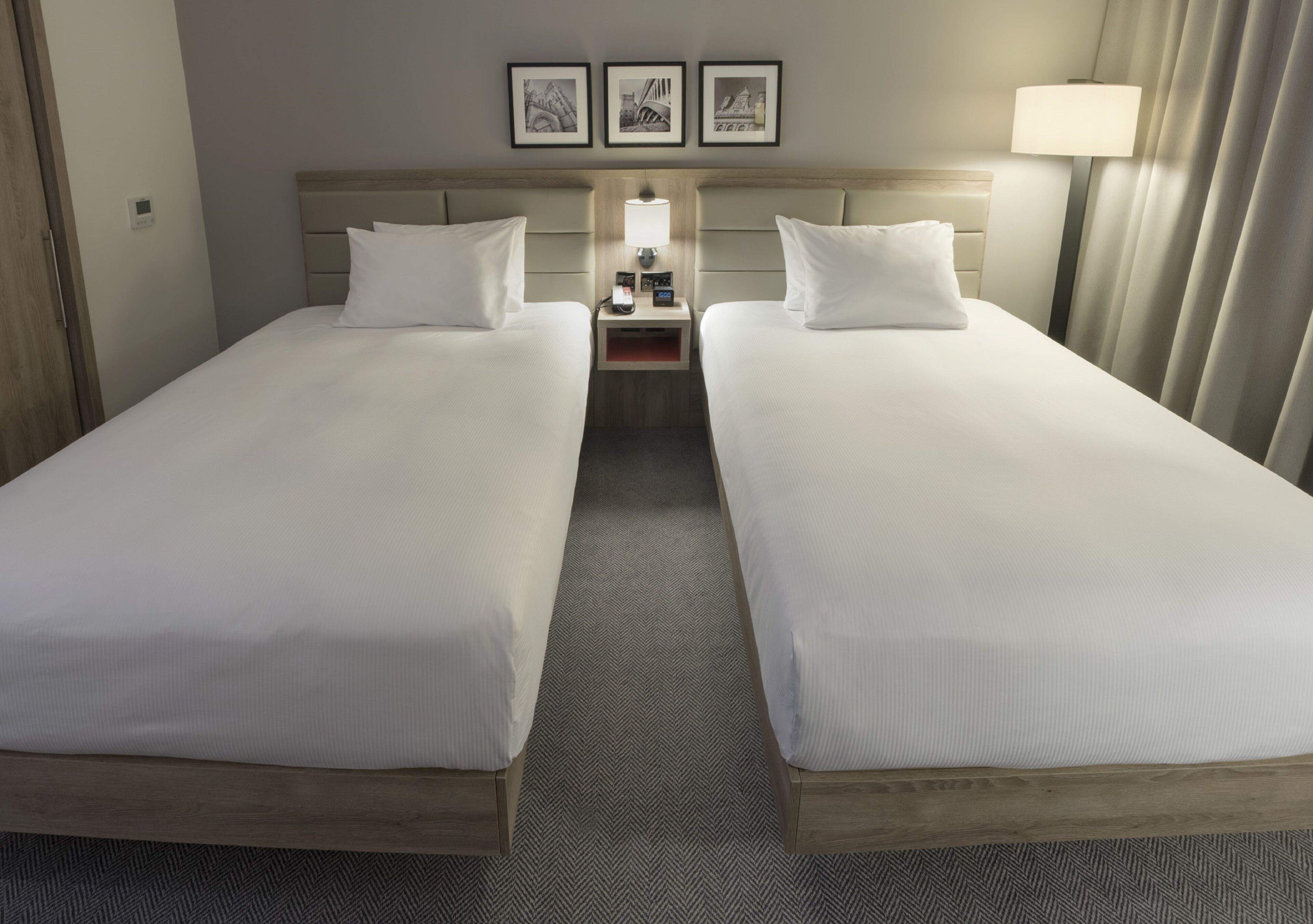 Quarto de hotel com duas camas de solteiro, uma mesa de cabeceira com abajur e três quadros na parede.